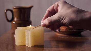 Close-up of a yellow candle in the shape of a star of David, her hand lights it. In the background, traditional food donuts and a cup. video