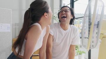 Two young Asian startup coworkers are fun blowing wind with electric fan, working in hot temperatures in a casual small business office in summer, refreshing from uncomfortable heat weather season. video