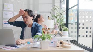 Two Asian workers are unhappy working with hot temperatures at casual office in summer season, uncomfortable with no air conditioner, using fans and drinking water, sitting at desks, and sweating. video