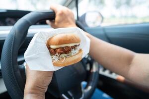 asiático dama participación hamburguesa a comer en auto, peligroso y riesgo un accidente. foto