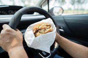 Asian lady holding hamburger and French fries to eat in car, dangerous and risk an accident. photo