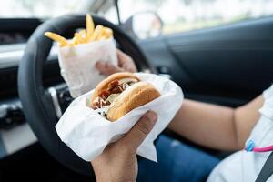 asiático dama participación hamburguesa y francés papas fritas a comer en auto, peligroso y riesgo un accidente. foto