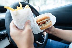 Asian lady holding hamburger and French fries to eat in car, dangerous and risk an accident. photo