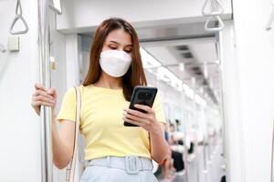 Concept of traffic and transportation. Asian female passenger wearing a mask holding a smartphone chatting online via the Internet. She was standing in the skytrain commuting in the city. photo