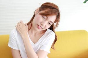 A beautiful Asian woman with pain in her right shoulder sits on the sofa in the living room. The concept of shoulder pain in adults. Office Syndrome. Health care. photo