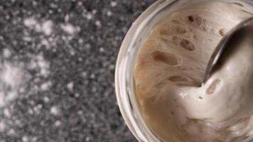 Close-up top view of a spoon stirring sourdough wheat leaven in a jar on a table with copy space. High quality FullHD footage video