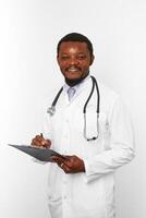 sonriente médico barbudo negro con túnica blanca con estetoscopio llenando registros médicos en el portapapeles foto