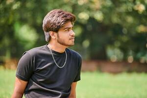 Attractive young indian man portrait in black t shirt and silver neck chain sitting on green lawn photo