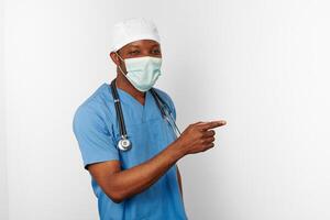 negro cirujano médico hombre en azul Saco blanco gorra y cirujano máscara señalando dedo a Derecha foto