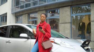 africano americano menina usando Smartphone e inclina-se em elétrico carro este é carregando. ambientalmente amigáveis transporte video