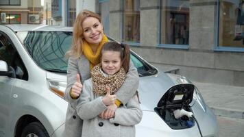 un mujer con su hija soportes cerca su eléctrico coche y mira a el cámara. cargando un eléctrico coche a un gas estación. ambientalmente simpático transporte video