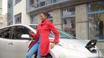 African american girl using smartphone and leans on electric car that is charging. Plug charging an electrical car. Electrical car recharging. Environmentally friendly transport video