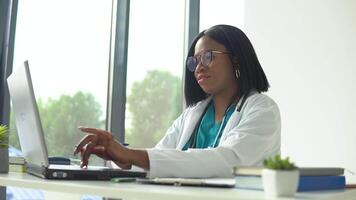 attraente africano femmina medico nel bianca cappotto e stetoscopio al di sopra di sua collo utilizzando il computer portatile a scrivania mentre Lavorando nel medico ufficio video