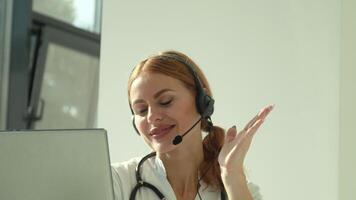 Professional female doctor in white medical coat and headset making conference call on laptop computer, consulting distance patient online in video chat
