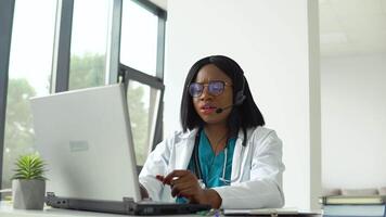 Professional african american female doctor in white medical coat and headset making conference call on laptop computer, consulting distance patient online in video chat