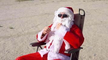 Père Noël claus dans des lunettes de soleil est assis sur une Soleil fainéant sur le Lac plage. Voyage et vacances concept video