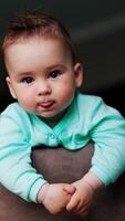 encantador niñito mirando alrededor en pie pacíficamente en el silla. adorable bebé demostración lengua y sonriente. bajo ángulo vista. vertical vídeo video