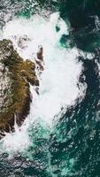 Craggy ragged rock in the blue ocean. White waves splashing by the cliff covered with sea salt. Top view. Vertical video