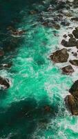 Turquoise water splashing by the stones at the shore. Rocky coastline of California with white waves splashing by. Vertical video