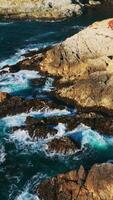 rojo liquen cubierta el harapiento rocas de Pacífico línea costera. blanco espumoso olas chapoteo por el peñascoso apuntalar de California. vertical vídeo video