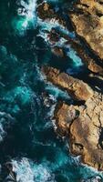 Brown rugged rocks covered with purple lichen at coastline. White waves splashing by the mountainous shore. View from above. Vertical video