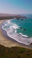 bellissimo ipnotizzante schiumoso onde in arrivo per il sabbioso spiaggia. roccioso riva coperto con erba. alcuni pietre fa capolino a partire dal acqua. aereo Visualizza. verticale video