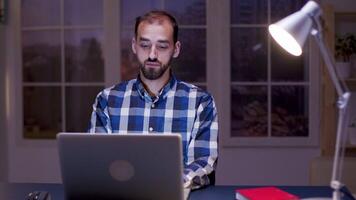 Thoughtful businessman typing on laptop while working late from home. Bearded businessman wearing a shirt and working from home at night. video