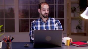 Bearded entrepreneur taking a sip of coffee and working on computer in his home office during night hours. video