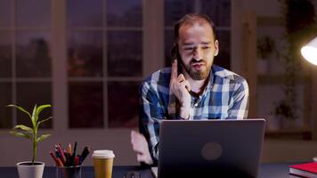 Bearded businessman having a conversation on his phone while working during night hours in his home office. video