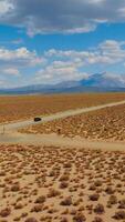 Yellow dry deserted landscape contrasting with bright azure sky with multiple clouds. Black car riding by the road. Mountains at backdrop. Vertical video