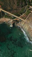 Flight above the highway along the mountains of California coastline. Arched bridge connecting the roads. Top view. Vertical video