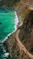 surpreendente cenário do escarpado pedras com rodovia em eles às costa do Califórnia. zangão vôo sobre a estrada com rochoso galpão em isto. lindo turquesa água com branco ondas perto pedras. vertical vídeo video