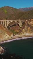 Beautiful sight of rocky shore with thin line of a sandy beach at Pacific coast. Arched bridge locating between two mountains with automobile road on it. Vertical video