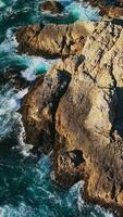 escabroso desnudo gris rocas a el línea costera de California en soleado día. blanco espumoso olas estrellarse por el acantilados aéreo vista. vertical vídeo video