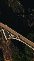 Descending above the gap between the mountains connected by the arched bridge. Cars run by the two-lane road on the bridge. Top view. Vertical video