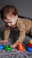 lief baby jongen kruipt omhoog naar een speelgoed- piramide Aan de vloer. zoet kind looks omhoog en gaat verder naar kruipen. wazig achtergrond. verticaal video