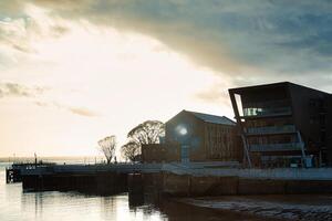 Serene sunset by the waterfront with silhouettes of buildings and trees, reflecting on calm water. photo