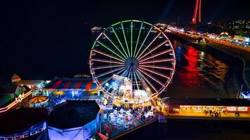 vistoso noche ver de un vibrante diversión parque con un ferris rueda y brillante luces reflejando en agua en piscina de fondo, Inglaterra. foto