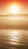 zon reflecterend in de zee water in de buurt de oever. silhouetten van bergen en sommige Kenmerken van stad nauwelijks gezien in de stralen van zonsondergang. verticaal video