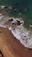 Thin line of sandy beach with some stones peeping from water. Beautiful white waves splashing to the shore on sunny day. Top view. Vertical video
