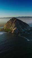 Magical sight of Morro Bay covered with thick fog. Triangle rock at the shore of Central coast of California, USA . Azure sky at backdrop. Vertical video