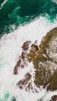 White foamy waves of aquamarine water splashing by the rock covered with salt. Brown algae accumulation on top of the ocean at California coast. Top view. Vertical video