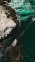 Rock covered with salt peeping out of aquamarine water of Pacific Ocean at California coastline. Algae floating on the surface of water. Aerial view. Vertical video