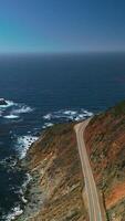 Rocky shore with highway over cliffs. Beautiful endless blue Pacific Ocean interflowing with clear azure sky. Aerial view. Vertical video