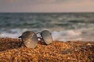 Side view of sunglasses on the beach near the turquoise sea with sand. Summer, beach holiday, stylish sunglasses, no people photo