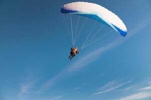 A paraglider takes off from a mountainside with a blue and white canopy and the sun behind. A paraglider is a silhouette. The glider is sharp, with little wing movement. photo