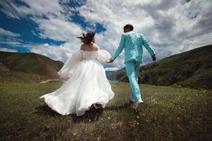 Rear view of running loving newlyweds holding hands, a bride in a wedding dress and a wife in a turquoise suit running across a field against the backdrop of green hills photo