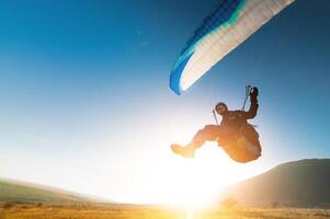 A paraglider takes off from a mountainside with a blue and white canopy and the sun behind. A paraglider is a silhouette. The glider is sharp, with little wing movement. photo