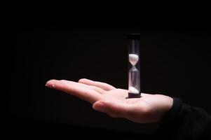 Close-up of a woman holding an hourglass in the dark. Fingers hold on both sides a plastic sand clock with white sand on a black background photo