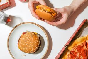 Hands holding a hamburger on a white table with pizza and drink photo